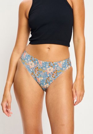Frau trägt hochgeschnittene Unterwäsche mit Blumenmuster in Blau-, Rosa- und Weißtönen, kombiniert mit einem ärmellosen schwarzen Croptop.