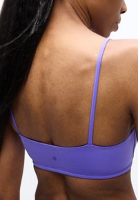 Η πλάτη μιας γυναίκας που φοράει μοβ αθλητικό σουτιέν με λεπτές τιράντες, μακριά μαύρα μαλλιά που πέφτουν πάνω σε έναν ώμο, με φόντο ένα απλό λευκό φόντο.