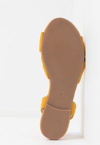 Keltainen nahkasaapas, jossa on tasainen, teksturoitu ruskea pohja, säädettävä remmi ja koko 37 merkintä pohjassa.