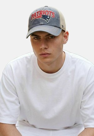 Young man wearing a gray and beige Patriots baseball cap and a plain white T-shirt, looking forward with a neutral expression.