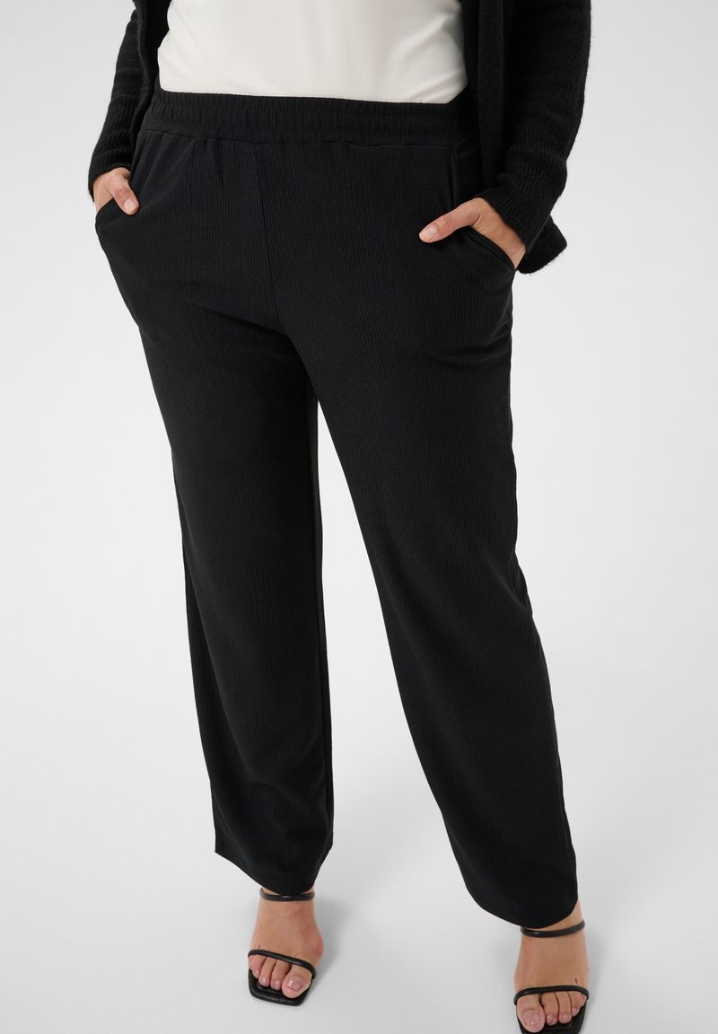 Pantalon noir ample avec taille élastique et poches latérales, porté avec des sandales noires à brides et un haut blanc.