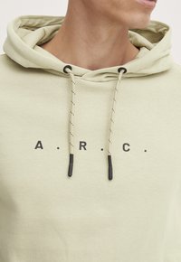 Lys grøn hoodie lavet af blødt stof. Har en frontlomme, snore med sorte accenter og trykte bogstaver "A.R.C." på brystet.
