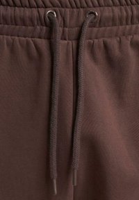 Un pantalon de survêtement marron présente une taille élastique avec un cordon de serrage. Le tissu est lisse avec une finition légèrement texturée.