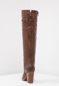 Bota de cuero marrón hasta la rodilla con punta redondeada, tacón ancho, cremallera lateral y un acento de hebilla en la parte superior del caña. Detalles de costura visibles.