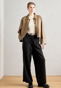 Braune Lederjacke, rippiertes beiges Oberteil, hochgeschnittene schwarze Hose, schwarze Plateau-Schuhe mit strukturierter Oberfläche und sichtbaren Knöpfen.