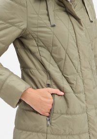 Olivgrüne, gesteppte Jacke mit einer Reißverschlusstasche. Mit Stehkragen und strukturiertem, gepolstertem Design. Weiches, leichtes Material.