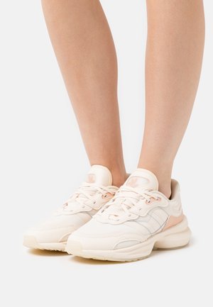 Jasno-beżowe buty sportowe z teksturowaną cholewką, białymi i brzoskwiniowymi akcentami, okrągłymi czubkami i płaskimi gumowymi podeszwami. Sznurowany fason z logo.