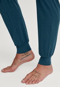Teal joggers met een zachte textuur en geplooide manchetten, gecombineerd met blote voeten met zichtbare tatoeages op de enkels en tenen.