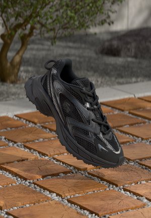 Zapato deportivo negro con materiales de malla y sintéticos, que presenta una superficie texturizada, punta redonda y diseño de cordones, colocado sobre una superficie pavimentada.