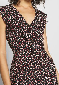 Vestido negro con escote en V y mangas con volantes, que presenta un patrón disperso de corazones rojos y beige. Tejido ligero con una textura suave.