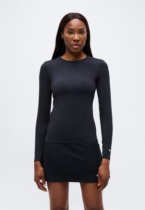 Woman wearing long-sleeve black athletic top and black sports skirt with Nike logos, standing against a plain light gray background.