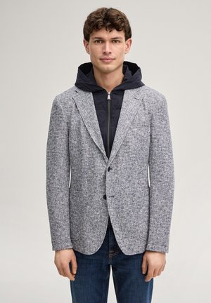 Junger Mann in einem grauen, strukturierten Blazer über einem schwarzen Kapuzenpullover mit Reißverschluss und blauen Jeans, der vor einem schlichten, hellen Hintergrund steht.