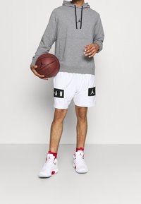 Hombre con sudadera gris, pantalones cortos de baloncesto blancos y zapatillas blancas sosteniendo un balón de baloncesto a la altura de la cintura frente a un fondo liso.
