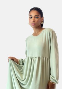 Hellgrünes Kleid aus weichem, strukturiertem Stoff mit lockerem Schnitt, langen Ärmeln und einem eng anliegenden Oberteil, das in einen gerafften Rock übergeht.