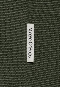 Olivgrüner gewebter Stoff mit strukturierter Oberfläche; beinhaltet ein weißes Etikett mit der Aufschrift "Marc O’Polo", das am Rand eingenäht ist.