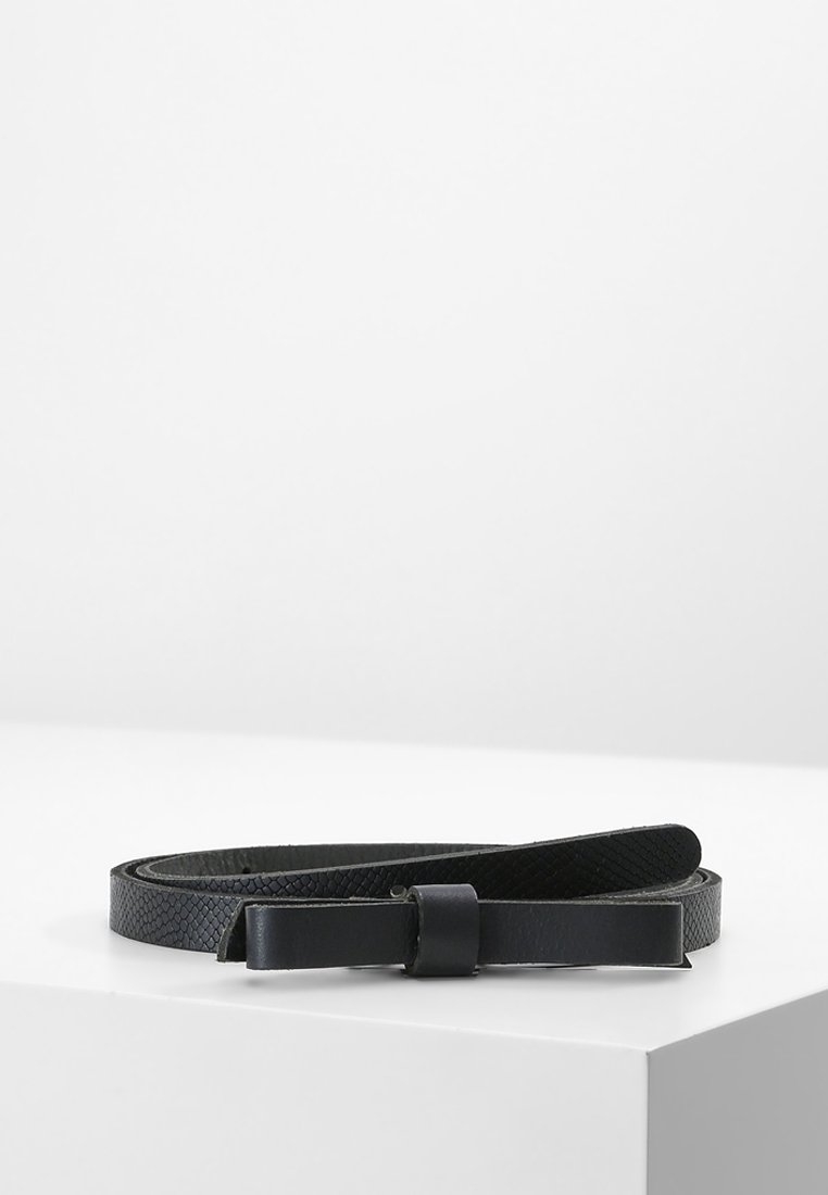 Ceinture en cuir noir texturé avec une boucle rectangulaire plate, enroulée sur une surface blanche inclinée, sur un fond clair uni.