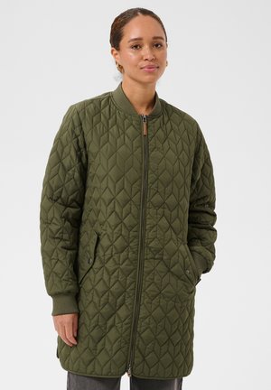Femme portant une longue veste matelassée vert olive avec une fermeture éclair à l'avant et des poches, debout, les mains dans les poches, sur un fond uni.