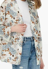 Giacca kimono floreale color crema, con fiori blu e marroni, apertura frontale, maniche lunghe e una vestibilità rilassata su un top bianco e jeans blu.