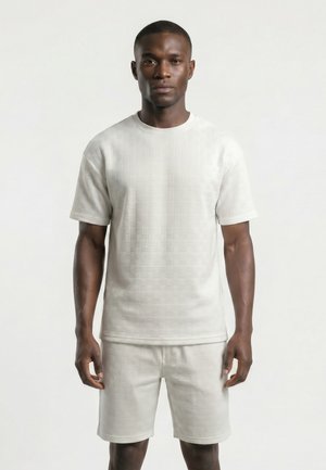 Homme portant une chemise blanche à manches courtes et un short assortis avec un motif tissé texturé, debout devant un fond clair uni.