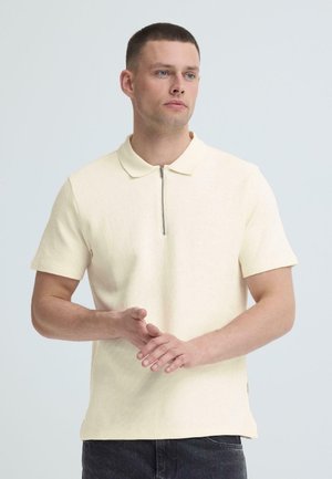 Vīrietis ar īsiem matiem, valkājot krēmkrāsas īsām piedurknēm adītu polo kreklu ar priekšā rāvējslēdzēju, stāv ar sakrustotām rokām priekšā.