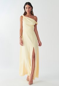 Gul klänning med en axel, tillverkad av lätt tyg, med en sidospalt och detaljerade ärmar med rynkning. Modellen bär sandaler med remmar.