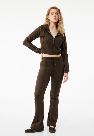 Młoda kobieta z długimi, kręconymi blond włosami, ubrana w ciemnobrązową krótką bluzę na zamek, dopasowane, rozszerzane spodnie i ciemne sneakersy, stojąca z ręką na biodrze.