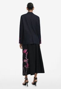 Tengerészkék blézer pink virágos hímzéssel a mandzsettákon, hosszú fekete szoknyával párosítva, amely pink virágmintákkal díszített. Fekete magas sarkú cipő egészíti ki az öltözetet.