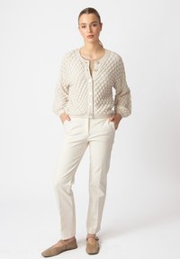 Bežani kardigan s teksturom i dizajnom mjehurića, sa dugmadi u obliku kruga, u kombinaciji s krem hlačama krojenim po mjeri i svijetlosmešnim cipelama.