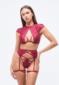 Σετ εσώρουχα από δαντέλα σε σκούρο κόκκινο χρώμα με τοπ από δαντέλα ψηλού λαιμού, σχέδιο με ανοιχτό μπούστο και πολύπλοκα λουριά στα ψηλόμεσα εσώρουχα με ιμάντες για ζαρτιέρες.