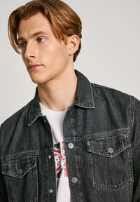 Zwarte denimjack met een kraag, knoopsluiting en twee borstzakken. Draagt over een wit graphic t-shirt.