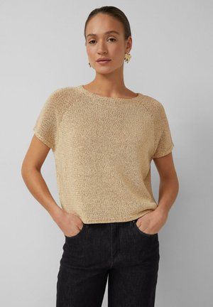 Vrouw in een beige gebreide top met korte mouwen en zwarte spijkerbroek, staand met handen in de zakken tegen een effen lichtgrijze achtergrond.
