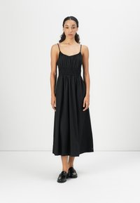 Abito lungo in cotone nero con spalline sottili, arricciato in vita, silhouette fluida e una texture liscia. Abbinato a scarpe nere.