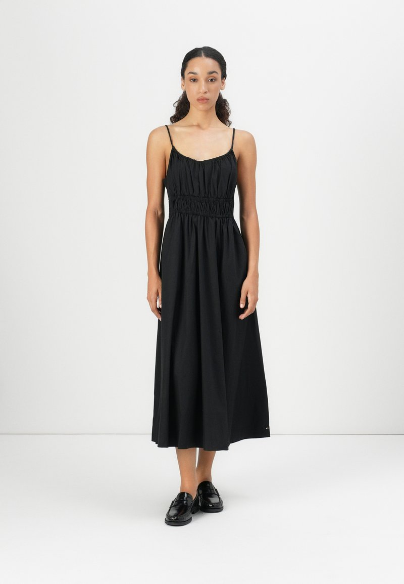 Abito lungo in cotone nero con spalline sottili, arricciato in vita, silhouette fluida e una texture liscia. Abbinato a scarpe nere.