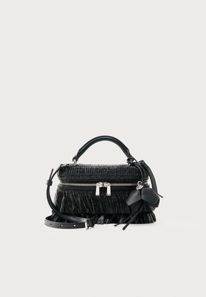 Μαύρη υφασμάτινη τσάντα χειρός με κορυφαία λαβή, ασημένιο φερμουάρ, ιμάντα ώμου, διακοσμητικές φούντες και γοητευτικό κρεμαστό σε σχήμα φιόγκου σε απλό ανοιχτό φόντο.