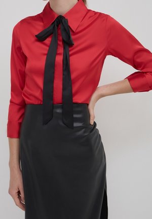Femme portant un chemisier rouge à manches longues avec un nœud papillon noir et une jupe en cuir noire taille haute, main reposant sur la hanche.