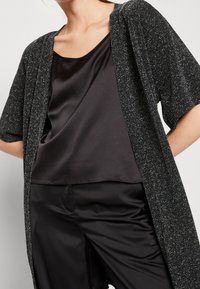 Svart skimrande kimono-kardigan med korta ärmar, som har ett lätt, strukturerat tyg och en öppen design, lager på lager över en svart satin-topp.