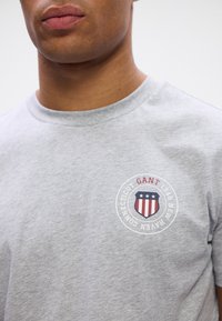 Grå bomulds t-shirt med et rundt logo med et skjolddesign, røde og blå striber, og teksten "GANT 1949 New Haven Connecticut."