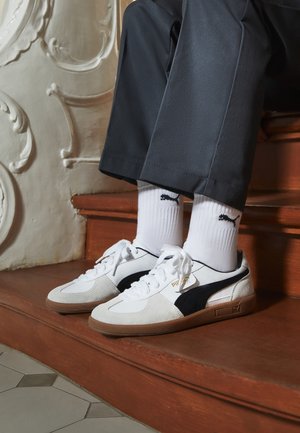Personne portant des baskets blanches Puma, des chaussettes blanches Puma et un pantalon foncé assise sur des escaliers en bois avec une décoration murale détaillée.