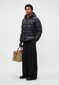 Doudoune noire avec capuche et design matelassé, associée à un pantalon large noir. Le modèle tient un sac marron avec des accents en cuir.