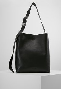 Czarna skórzana torba na ramię z regulowanym paskiem i subtelnym logo Calvin Klein wytłoczonym na przodzie, prezentowana na białym piedestale.