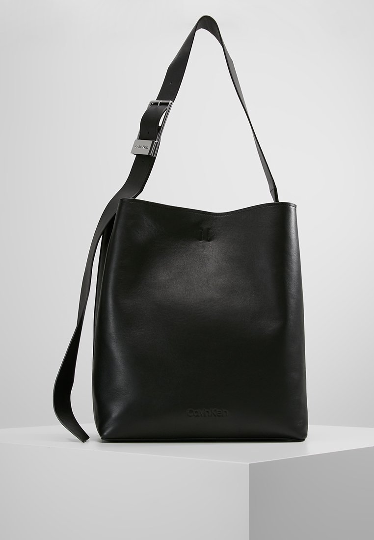 Czarna skórzana torba na ramię z regulowanym paskiem i subtelnym logo Calvin Klein wytłoczonym na przodzie, prezentowana na białym piedestale.