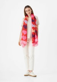 Écharpe rouge avec des motifs de cœurs multicolores, en tissu respirant, bordée d'un design rose. Portée sur une chemise claire et un pantalon blanc.