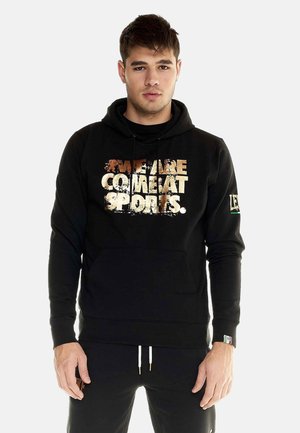 Jeune homme debout portant un sweat à capuche noir avec un texte usé affichant "WE ARE COMEAT SPORTS" sur le devant et un pantalon noir assorti.