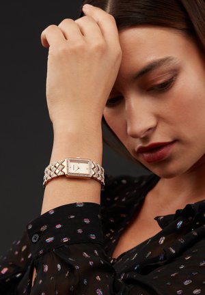 Rose gold watch with a rectangular face, diamond accents, and a woven band. Worn on a wrist with a dark, patterned blouse.