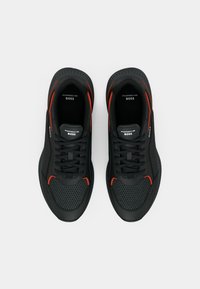 Baskets athlétiques noires avec panneaux en mesh et en matériau solide, présentant des accents rouges, des lacets ronds et un logo sur la languette et les côtés.