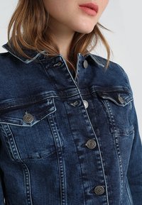 Mörkblå denimjacka med silverfärgad hårdvara, knappstängning, två bröstfickor och kontrasterande sömmar. Har en klassisk krage.