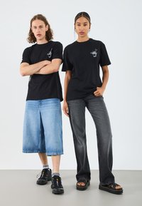 Sorte grafiske t-skjorter med et tegneserieuttrykk, kombinert med lyseblå denim culottes og mørkegrå rette jeans. Svarte sko med detaljer.