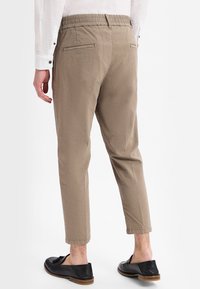 Man in beige cropped pants, een wit overhemd met lange mouwen en zwarte instappers, gezien vanaf de achterkant tegen een witte achtergrond.