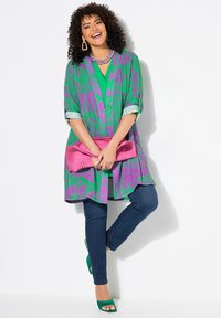 Bunte lange Bluse in Grün mit lila Blumenmustern, kombiniert mit einem türkisfarbenen Oberteil und dunklen Jeans. Die Accessoires umfassen eine pinke Clutch und grüne High Heels.