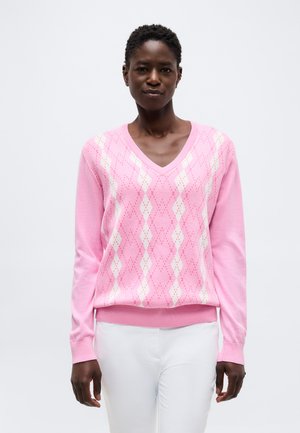 Femme portant un pull à motif losanges rose à col en V et un pantalon blanc, debout devant un fond clair uni.